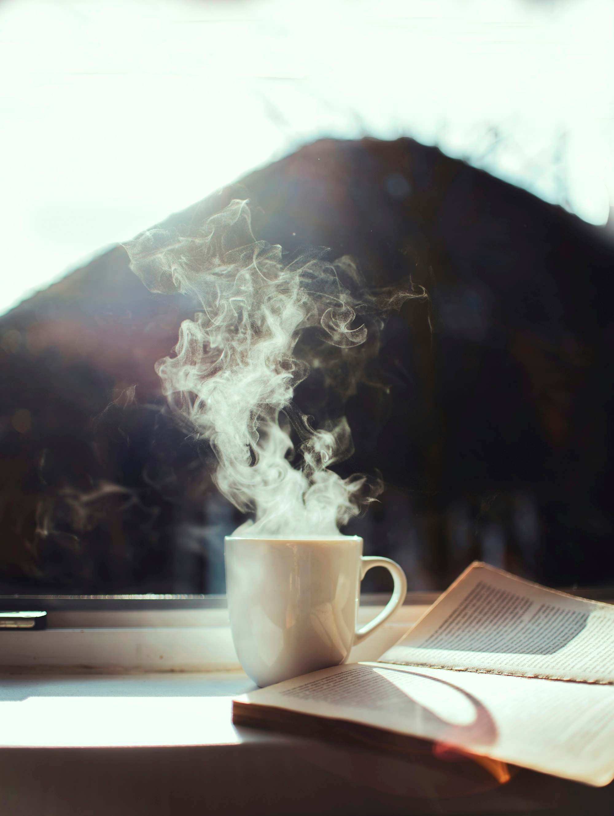 Taza de café humeante junto a un libro abierto en el marco de una ventana con luz de mañana
